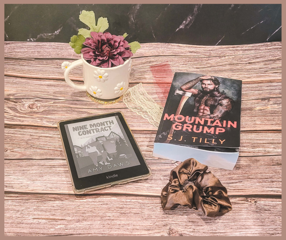 A wooden table with a Kindle showing "Nine Month Contract" by Amy Daws, a book titled "Mountain Grump" by S.J. Tilly, a flower mug, and a scrunchie.
