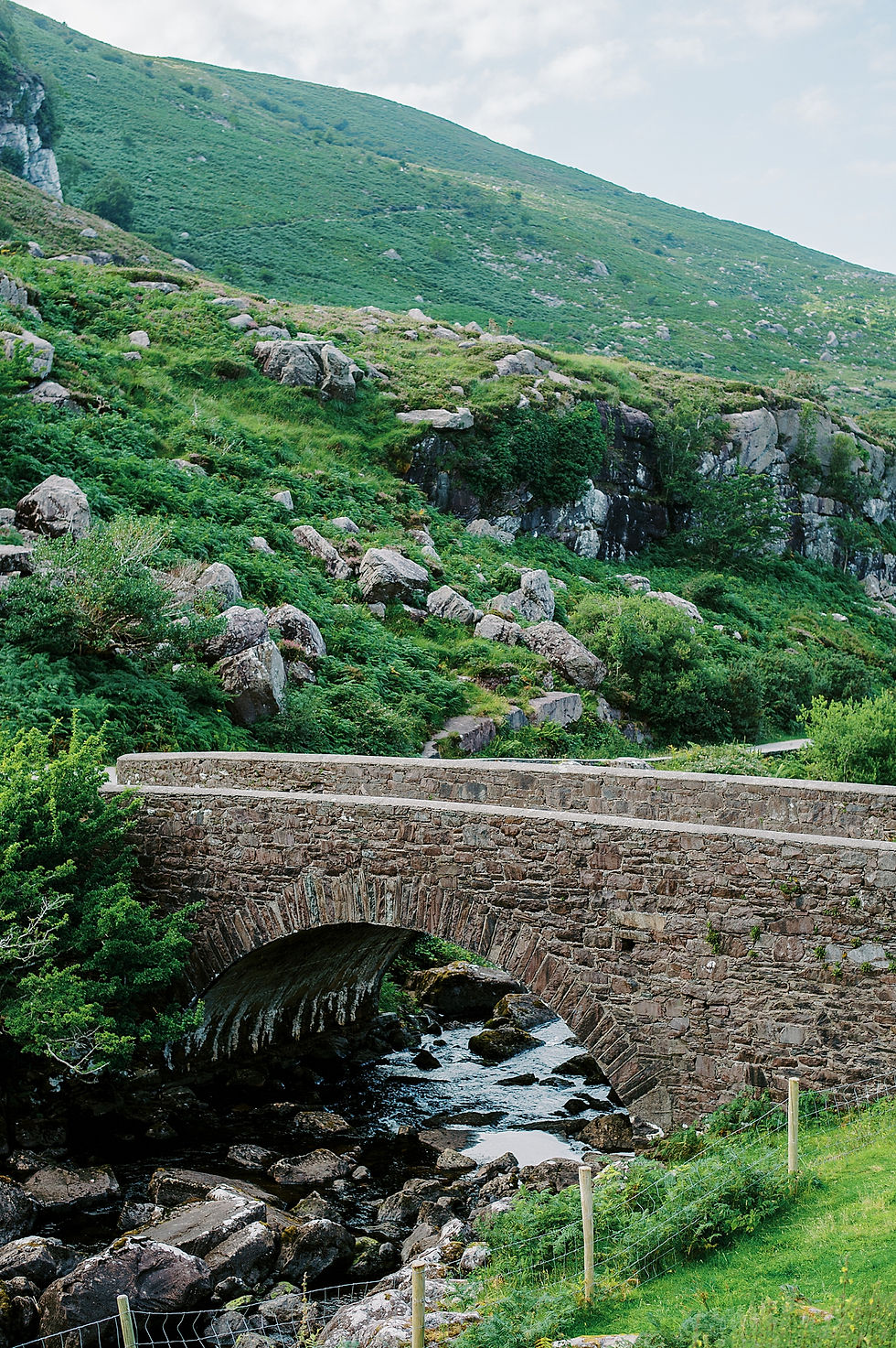 Stone arch bridge over a rocky stream in a lush green hillside, with mossy rocks and cloudy sky.