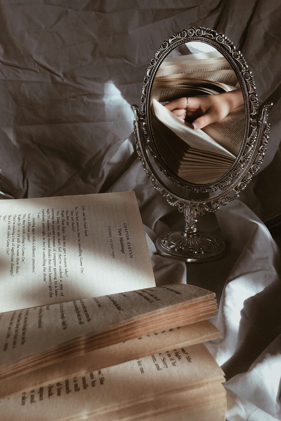 An ornate mirror reflects a hand turning pages of a book on a crumpled fabric surface. The open book reveals text, creating a serene mood.