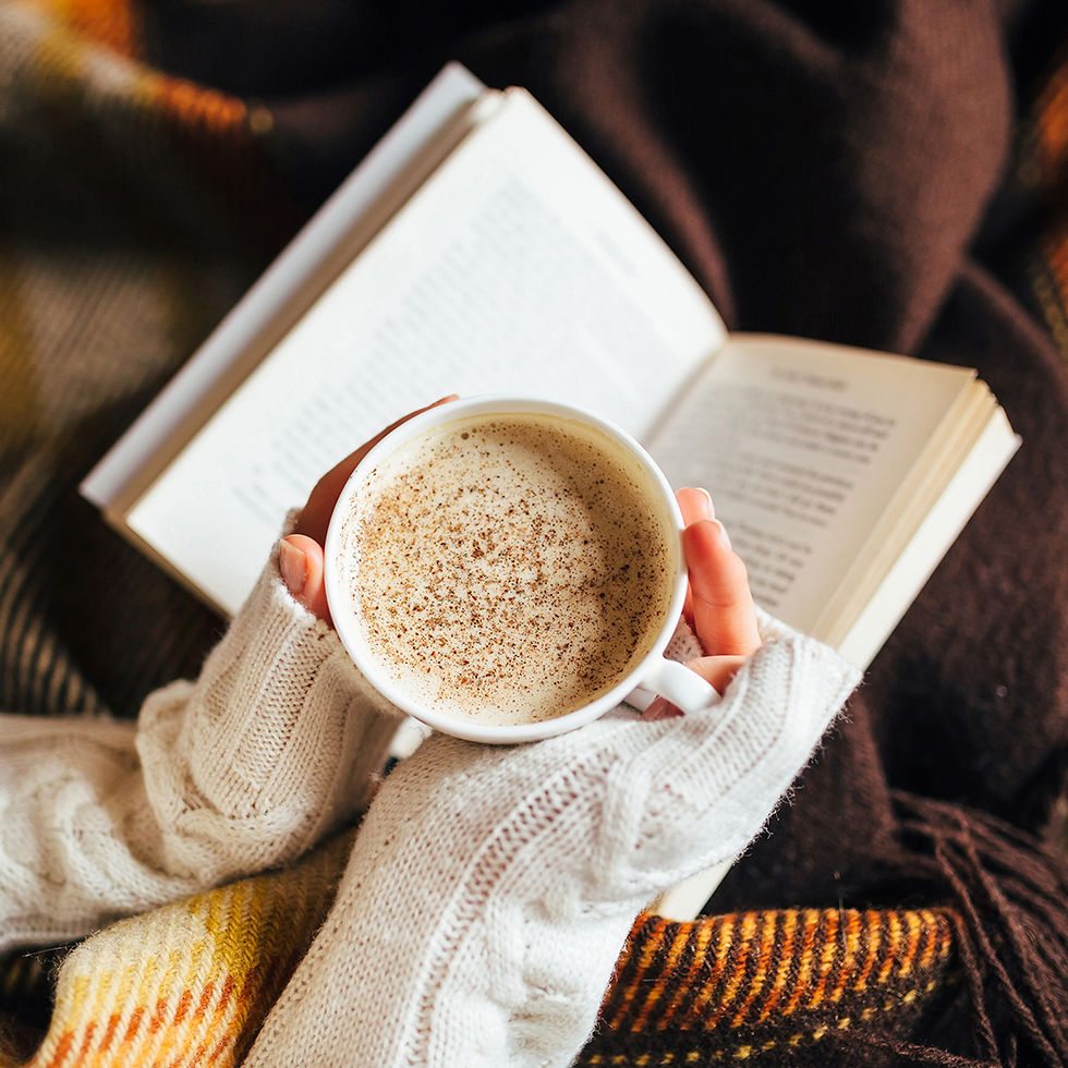 Hands in cozy sweaters hold a spiced latte over an open book. Brown and yellow plaid blanket background creates a warm, relaxed vibe.