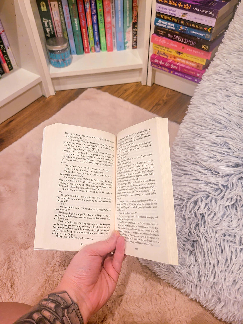 A tattooed hand holds an open book on a fluffy beige rug. Bookshelves with colorful books are visible in the background. Cozy setting.