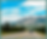Car driving on a mountain highway beneath bright clouds, with pine forest and rocky peaks under a blue sky.