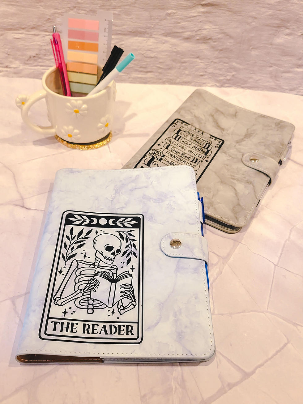 Notebooks with skeleton and book designs are on a light marble surface. A daisy-patterned mug holds pens and sticky notes.