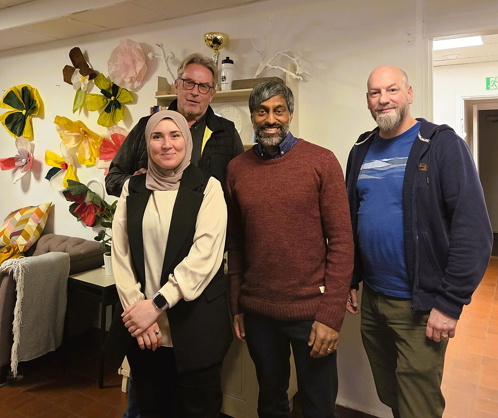 Fyra personer, en kvinna och tre män, poserar för kameran. I rummet de står syns en soffa och väggdekorationer och bakgrunden.