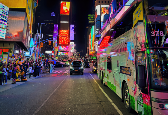Tour bus, traffic, and sidewalk crowds beneath colorful Times Square billboards at night.
