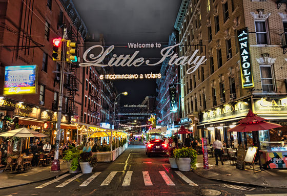 Illuminated restaurants and diners beneath the Welcome to Little Italy street sign at night.