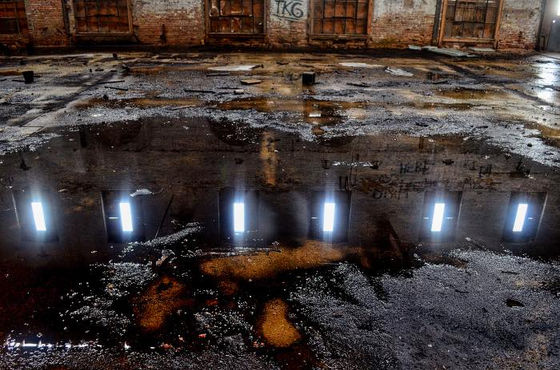 Bright roof openings reflected in a broad puddle on a damaged industrial floor.