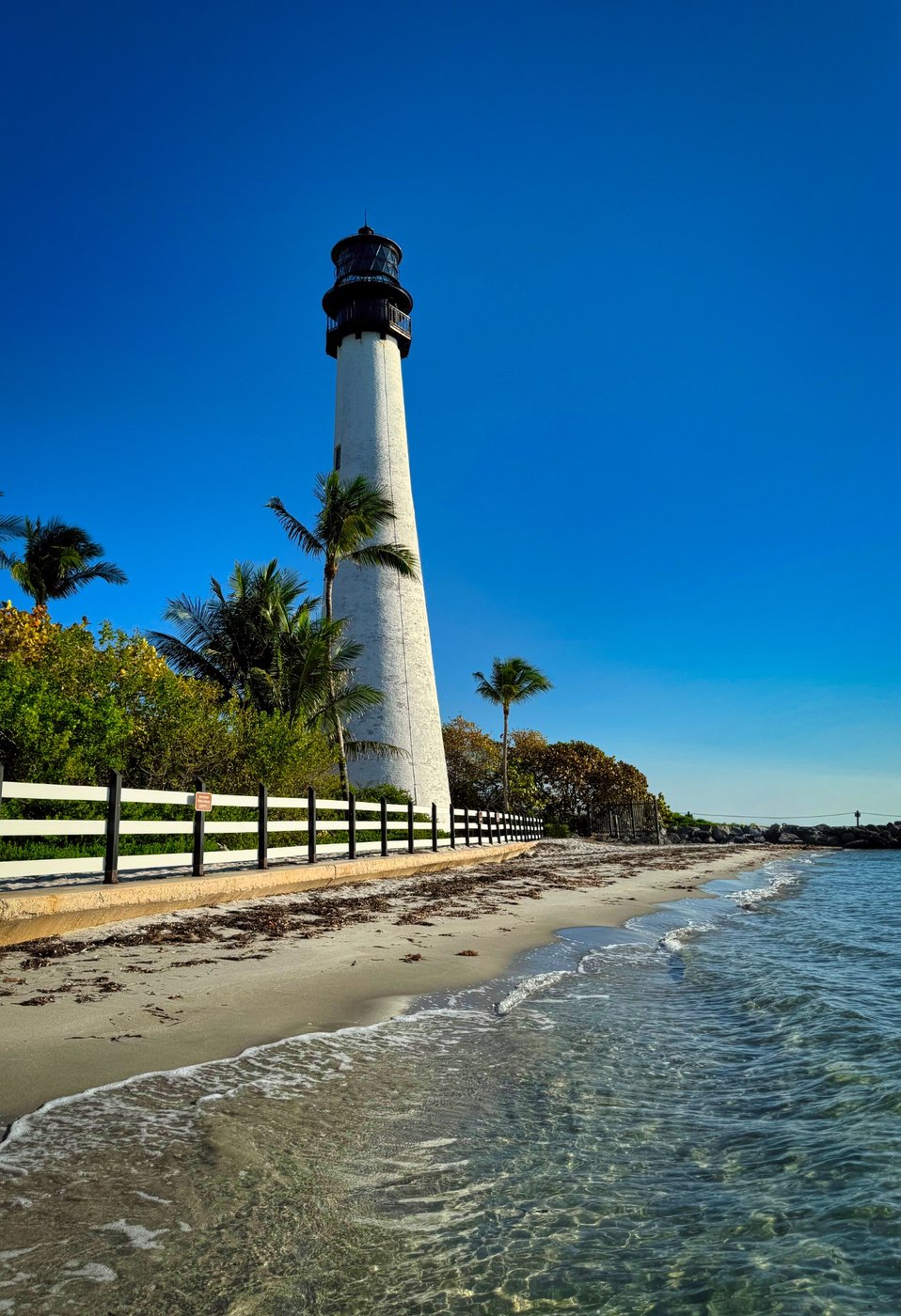 Historic Beacon of the Tropical Coastline White lighthouse beside palms, a narrow beach, and clear shallow water.