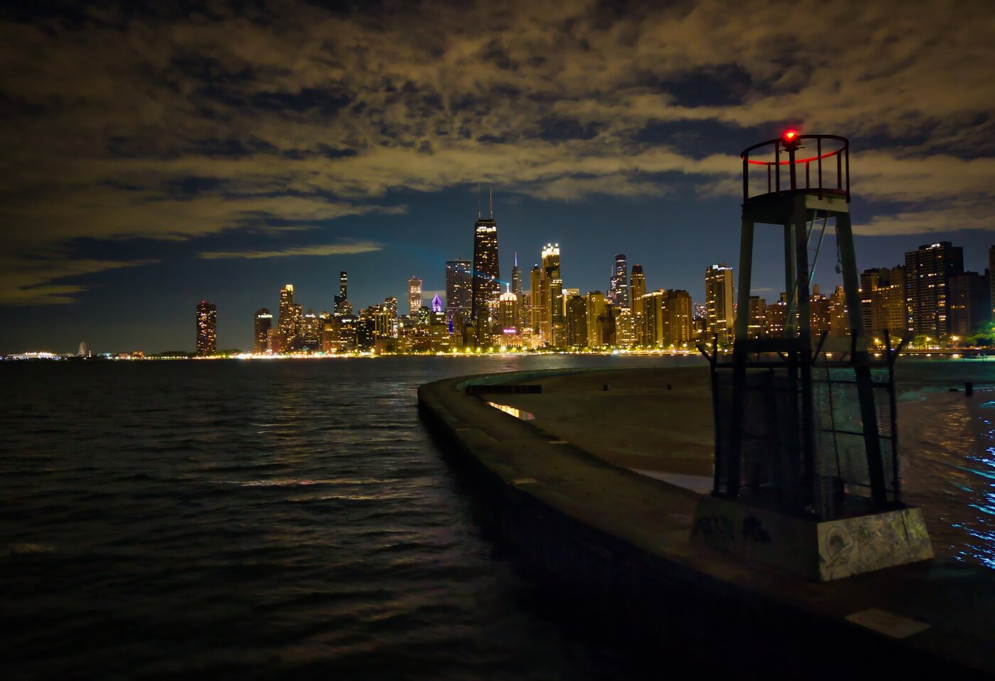Beacon on the Edge Dark beacon at the right edge of a curved pier with Chicago skyline lights across the water.