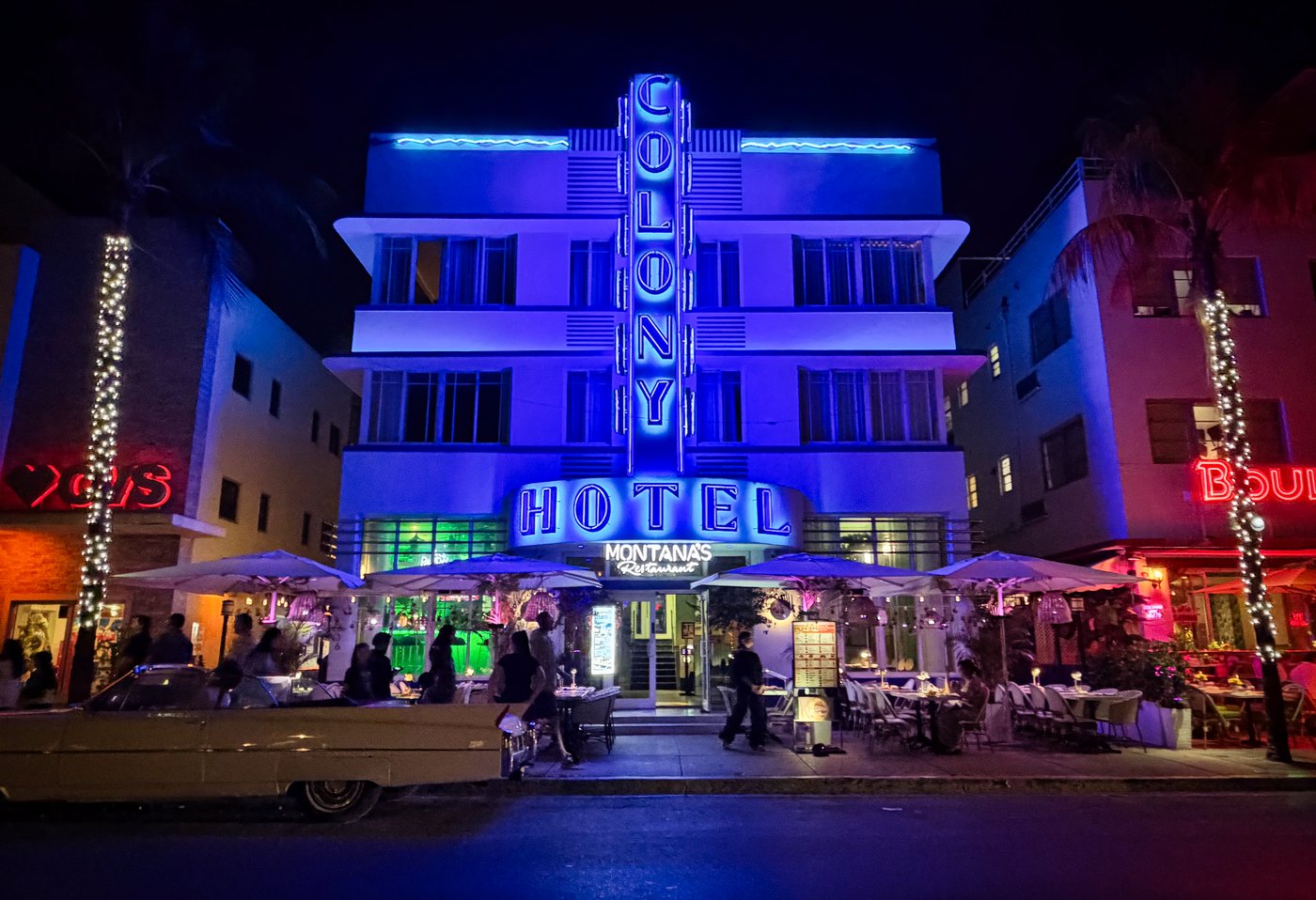 Colony in Cobalt Colony Hotel illuminated in blue above a busy nighttime sidewalk.