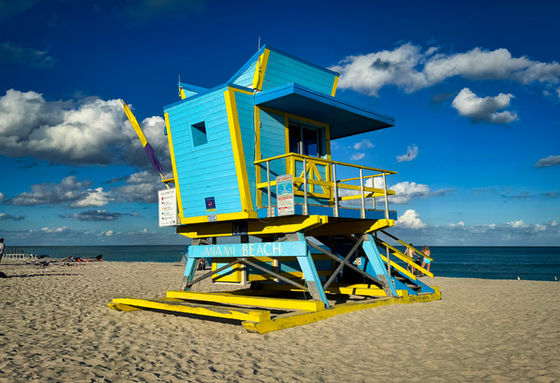 Turquoise and yellow lifeguard tower on sand beneath scattered clouds.