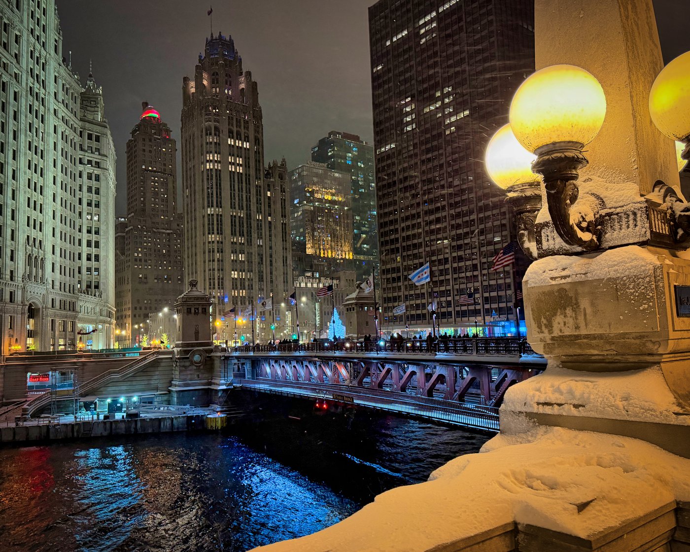Snow-covered Chicago riverfront lamps beside a bridge and illuminated skyscrapers at night.