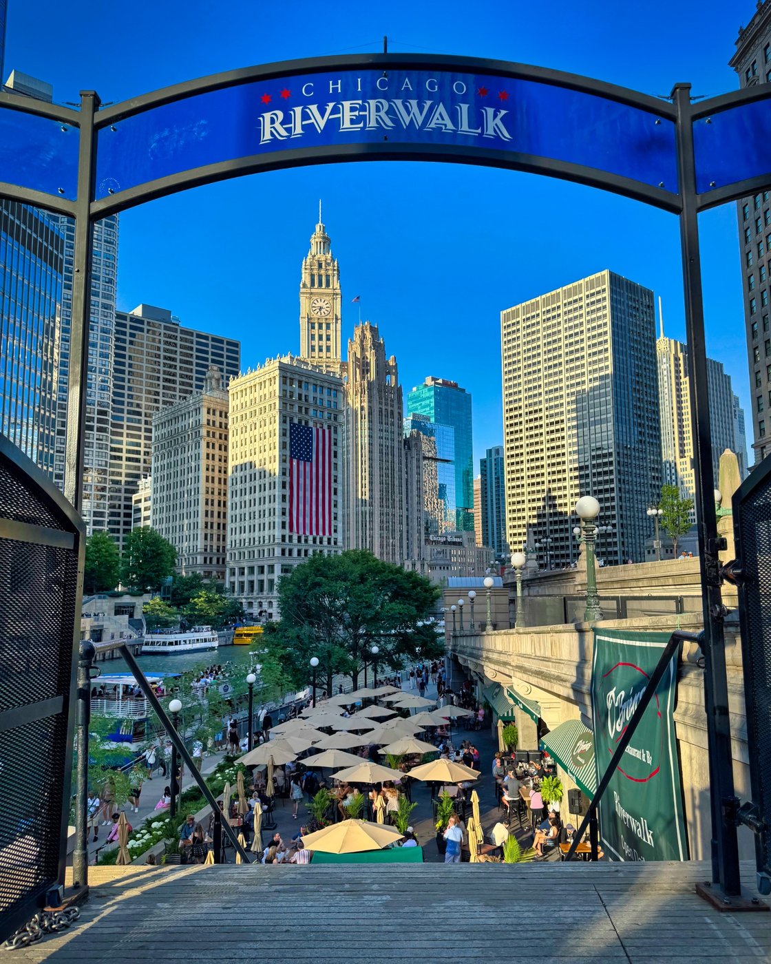 Chicago Riverwalk entrance arch above steps, umbrellas, and tall buildings.