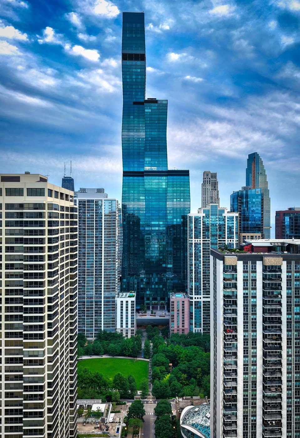 A Tower of Shifting Planes Tall offset blue-glass skyscraper rising above Chicago rooftops beneath textured clouds.
