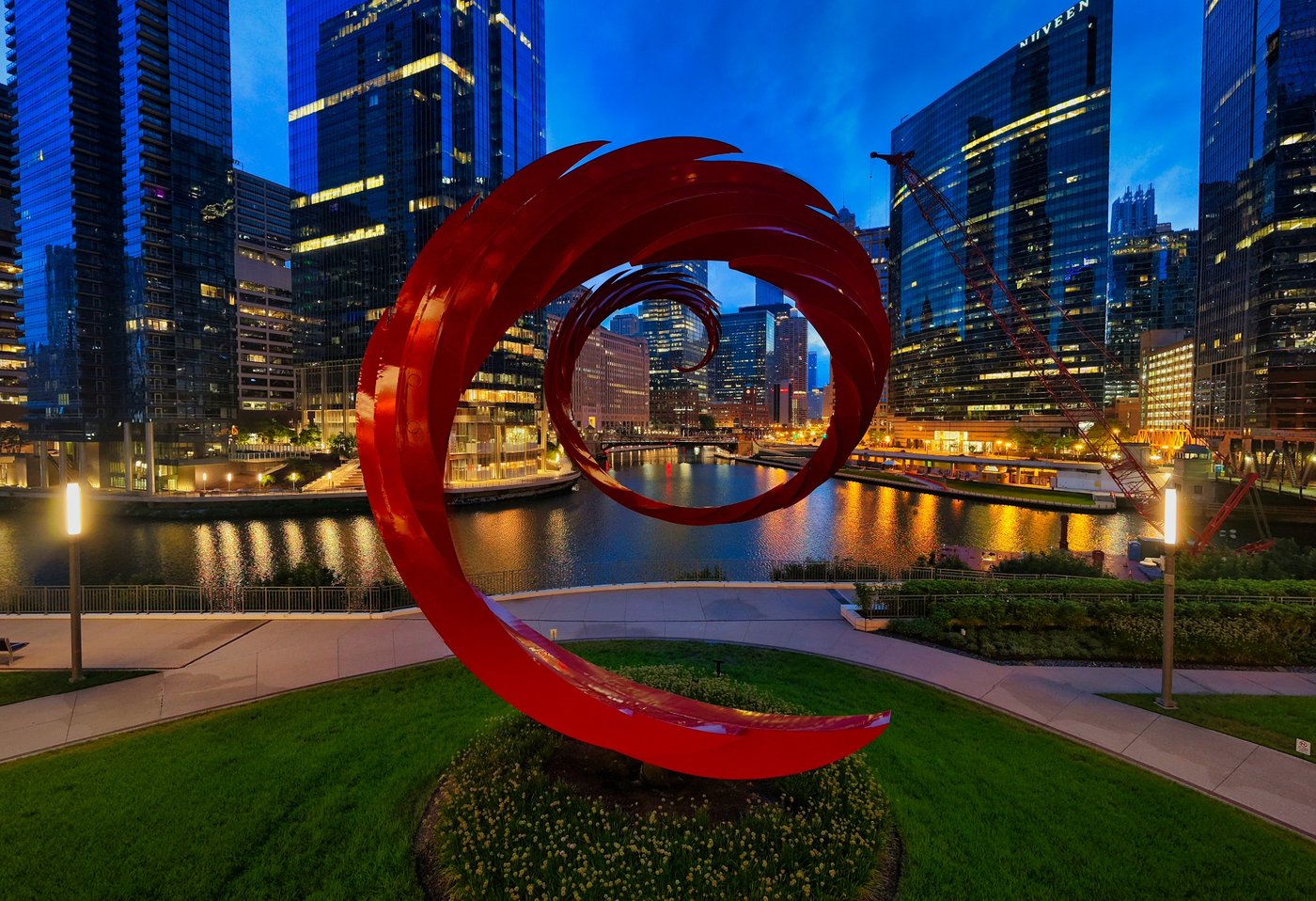 The Red Curve After Dark Red spiral sculpture beside a river and illuminated Chicago glass towers at night.