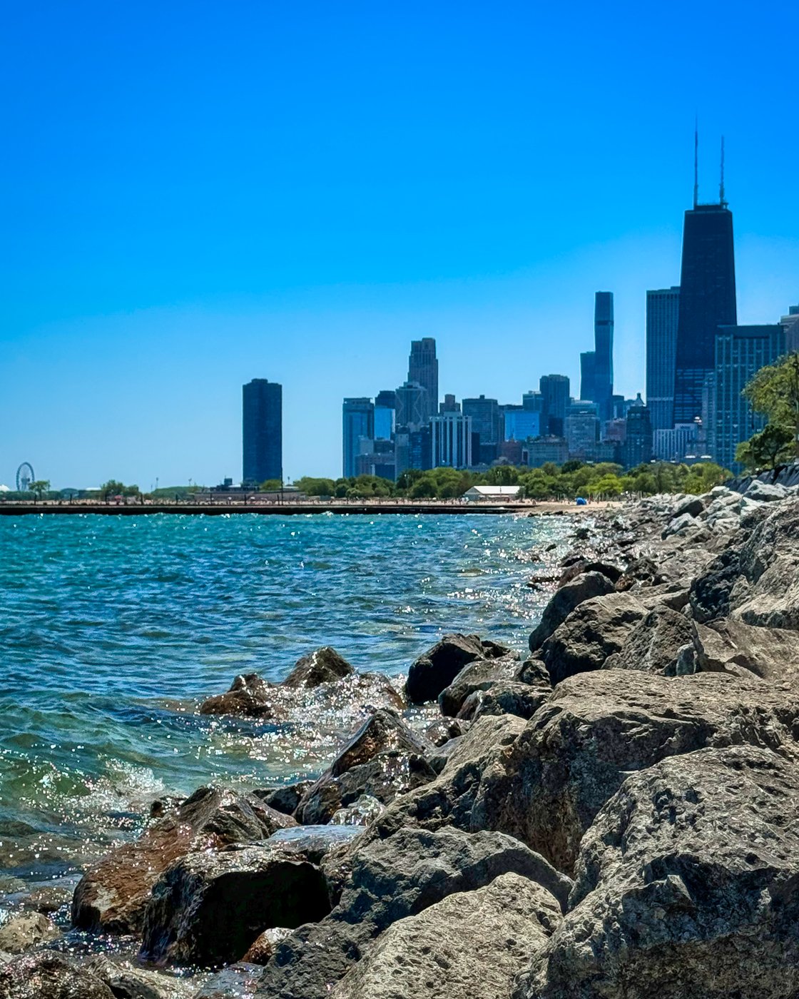Rocky Lake Michigan shoreline leading toward Chicago skyscrapers beneath blue sky.