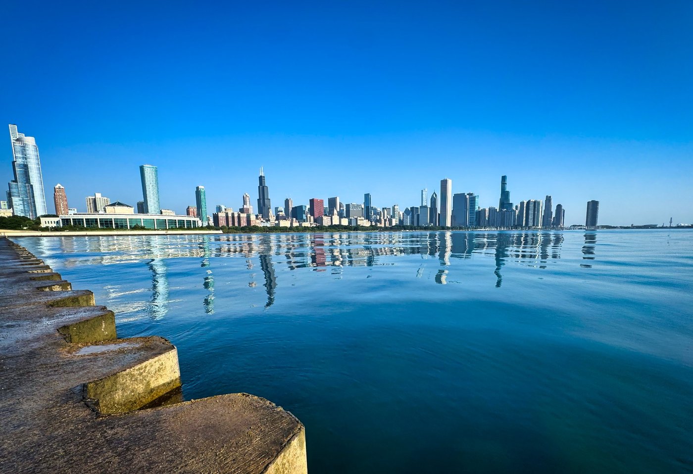 Morning Reflections On Lake Michigan Chicago skyline reflected in calm blue lake water beside stepped concrete shoreline.