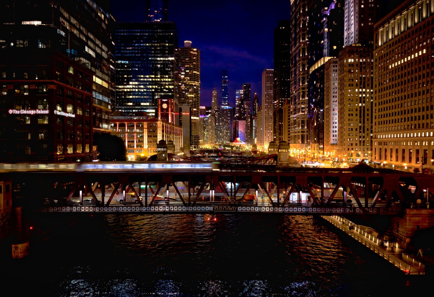 The River's Night Passage Nighttime Chicago River view across a steel bridge toward brightly illuminated buildings.