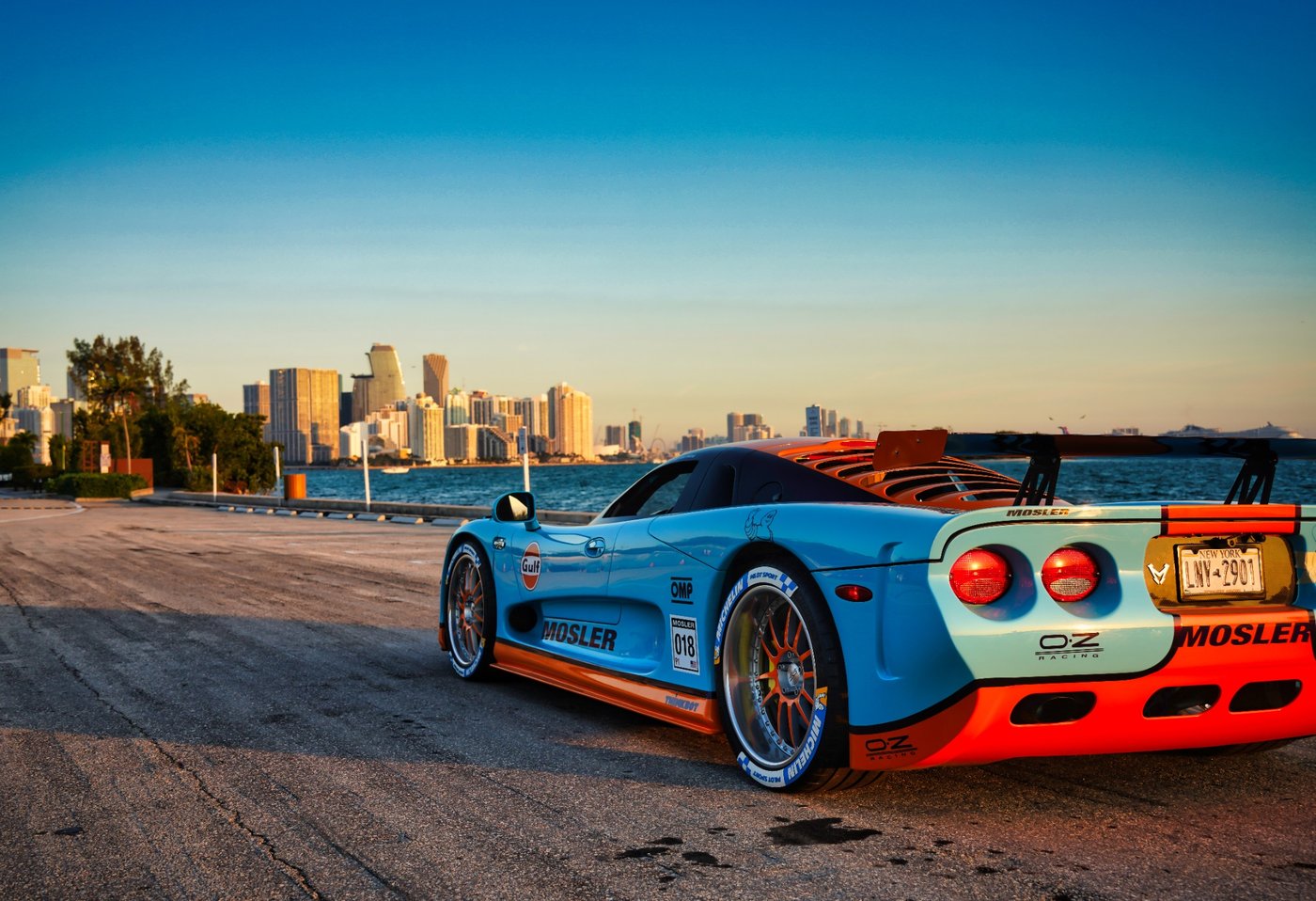 Sunset Velocity Blue and orange sports car on a waterfront road with a distant skyline.
