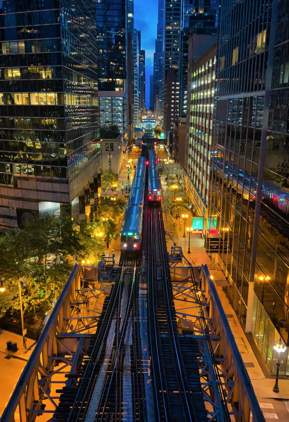 A Corridor of Light Elevated tracks and streetlights between tall Chicago buildings in a vertical evening view.