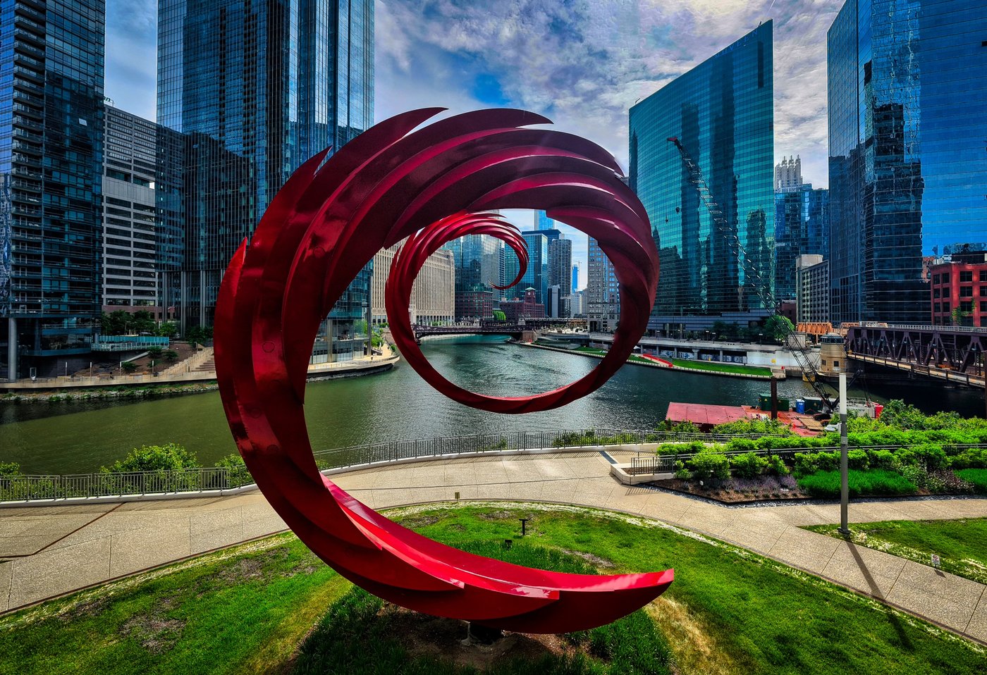 A Red Turn Red spiral sculpture in a riverside garden surrounded by Chicago skyscrapers in daylight.