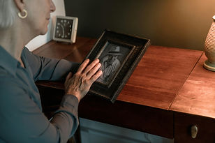 Een oudere vrouw strijkt met haar hand over een ingelijste zwart-wit foto op een houten bureau. Er is een klok en een lamp aanwezig.