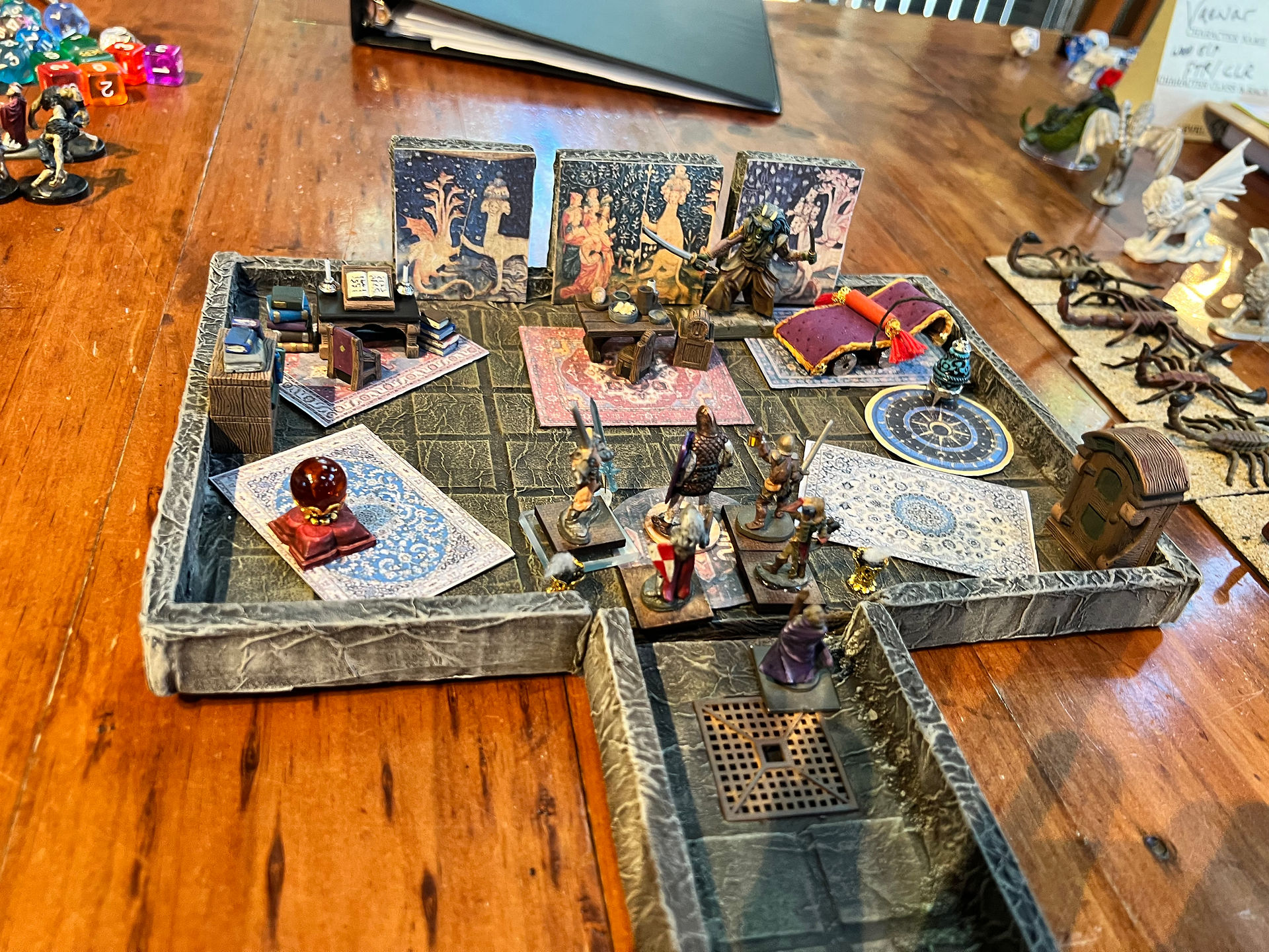 Photo of a tabletop dungeon room with painted miniatures exploring a decorated chamber. The room contains rugs, wall hangings, a fountain or basin, a bed, and other furnishings, with figures gathered near the center and entrance.