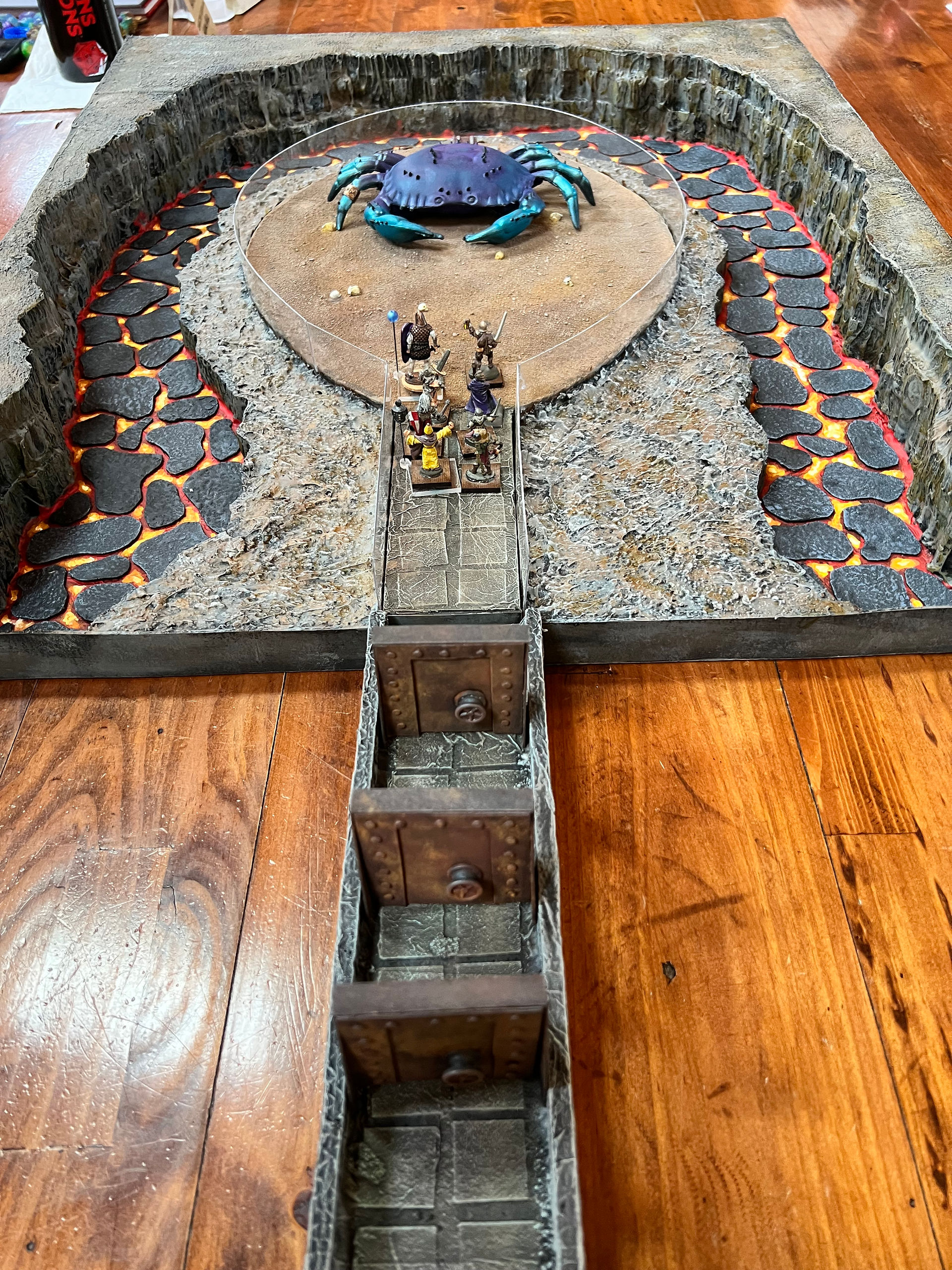 Photo of painted fantasy miniatures standing at the entrance to a stone platform or chamber, facing a large blue crab-like monster in the background. The scene appears to depict an adventuring party confronting a giant creature across a dungeon or cavern floor.