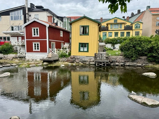 Red and yellow wooden houses line a calm canal, their reflections mirrored in the water under a cloudy sky.