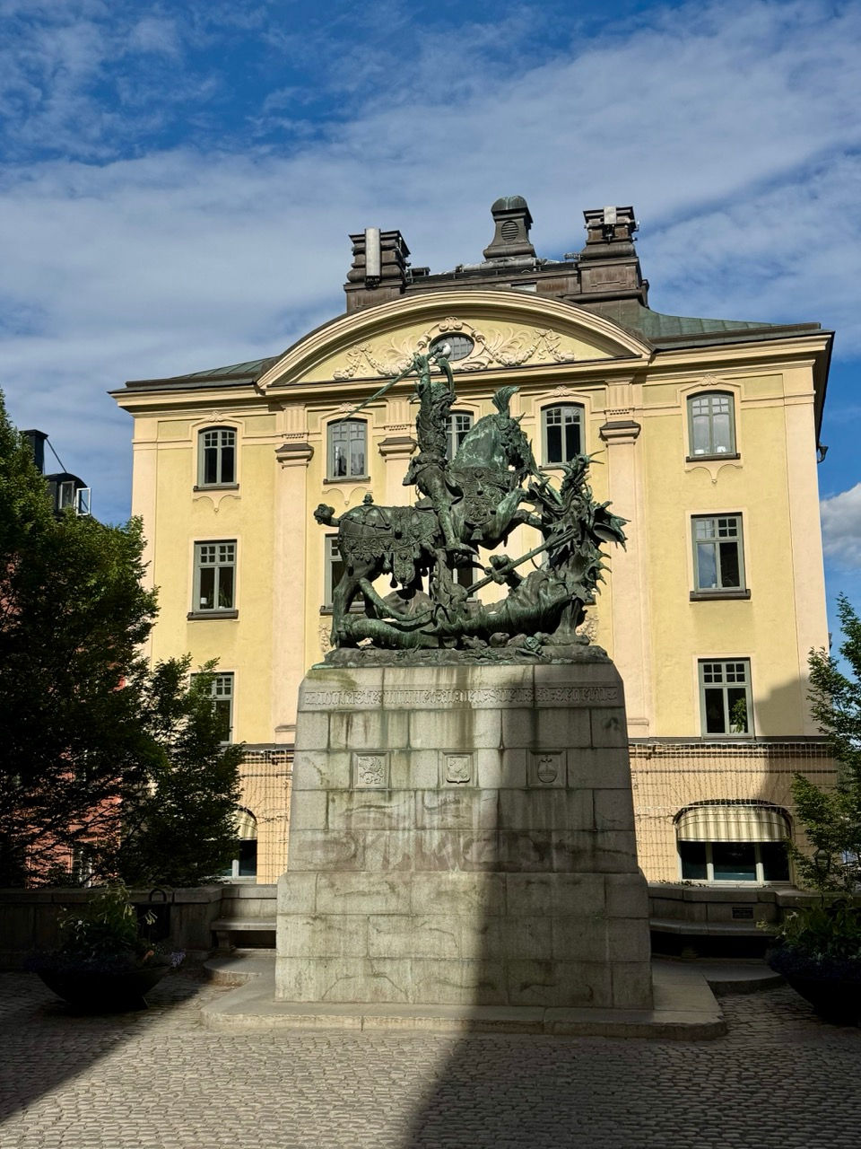 15th century bronze statue of Saint George and the Dragon before a pale yellow historic building under a blue, partly cloudy sky in a quiet courtyard