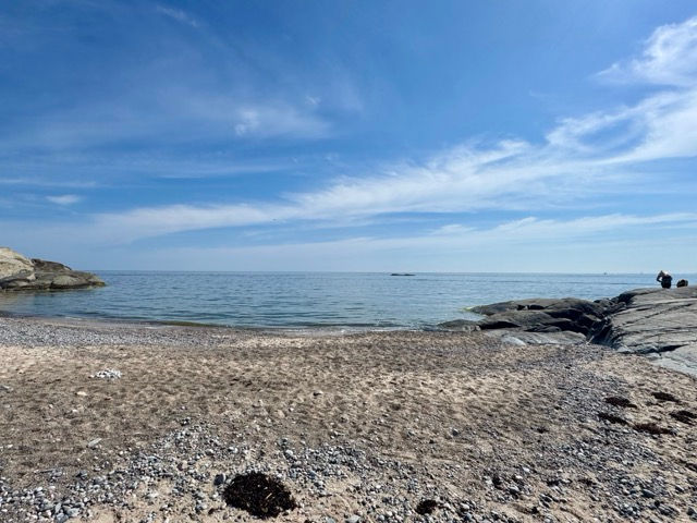 Rocky pebble beach by calm sea under a bright blue sky with wispy clouds; a person stands on the right by the rocks.