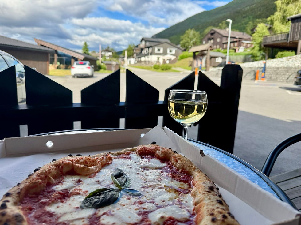 Pizza in a box beside a glass of white wine on an outdoor table, with a quiet mountain village and blue sky behind.