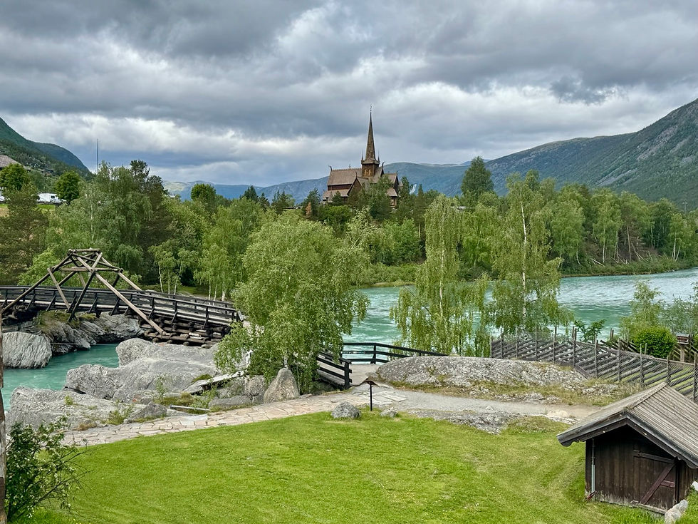 Wooden bridge by a turquoise river, with a stave church on a hill, green trees, mountains, and a cloudy sky.