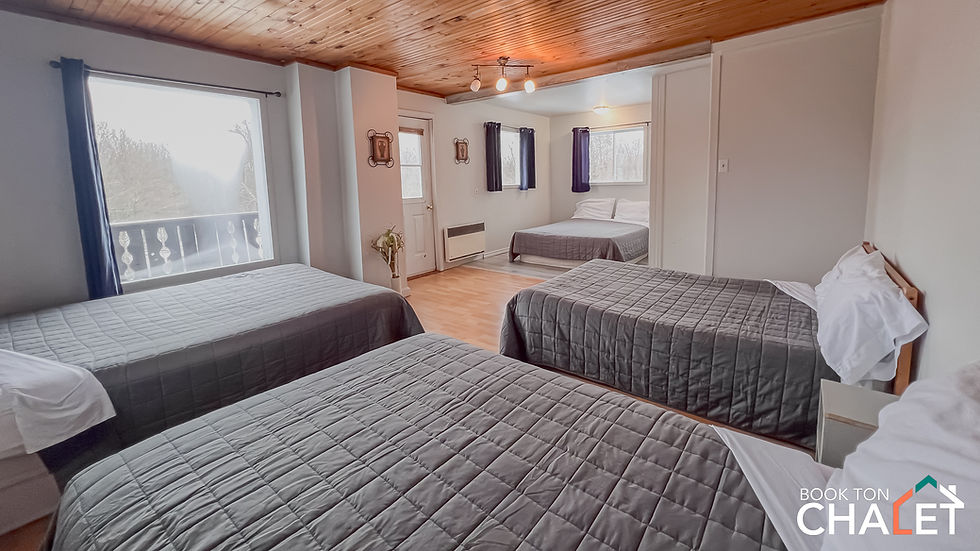 Chambre spacieuse et lumineuse avec quatre lits doubles recouverts de couvre-lits gris. Le plafond est en bois, ajoutant une touche chaleureuse à l’espace. De grandes fenêtres avec rideaux sombres laissent entrer la lumière naturelle et offrent une vue sur l’extérieur. Le plancher en bois clair complète le décor épuré. Idéal pour accueillir plusieurs personnes dans un confort simple et fonctionnel.