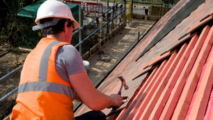 Image of a builder wearing a orange high vis jacket hammering a tile onto a roof with red battens