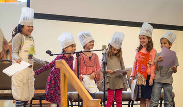 Children wearing chef hats and aprons stand on the stage of the UUCF sanctuary during Sunday Service to share a skit prepared in their Youth Exploration program on Sundays