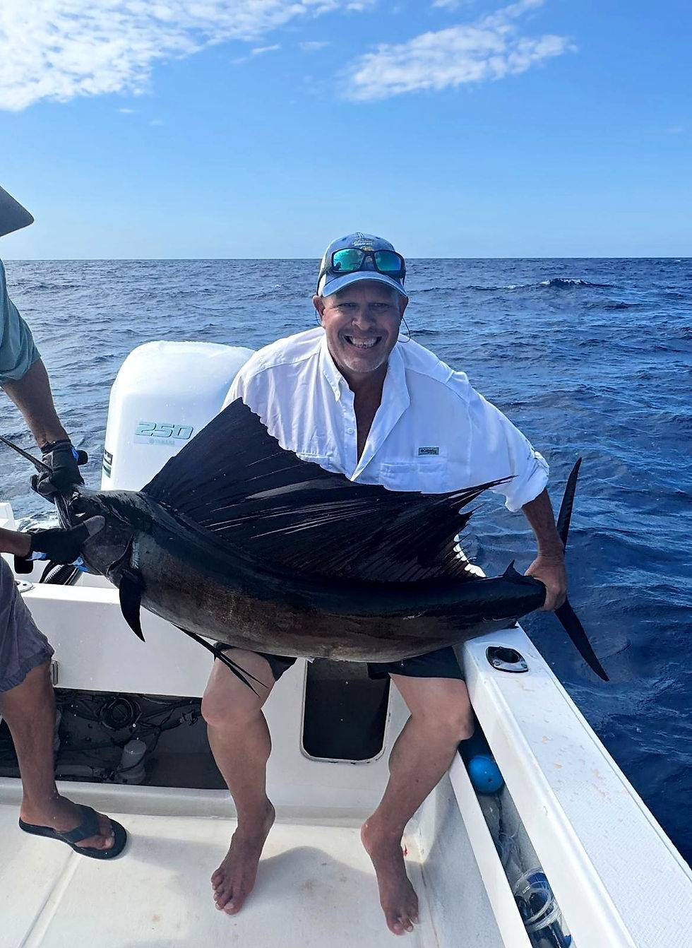 Smiling angler in a white fishing shirt holding a sailfish with its dorsal fin raised on a center console boat, deep blue Sea of Cortez behind