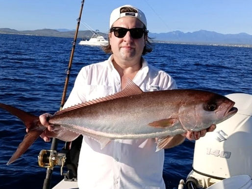 Angler in a white cap, sunglasses and white fishing shirt holding a large bronze amberjack on a center console boat with a Suzuki outboard, deep blue sea, a sportfishing boat and mountains behind