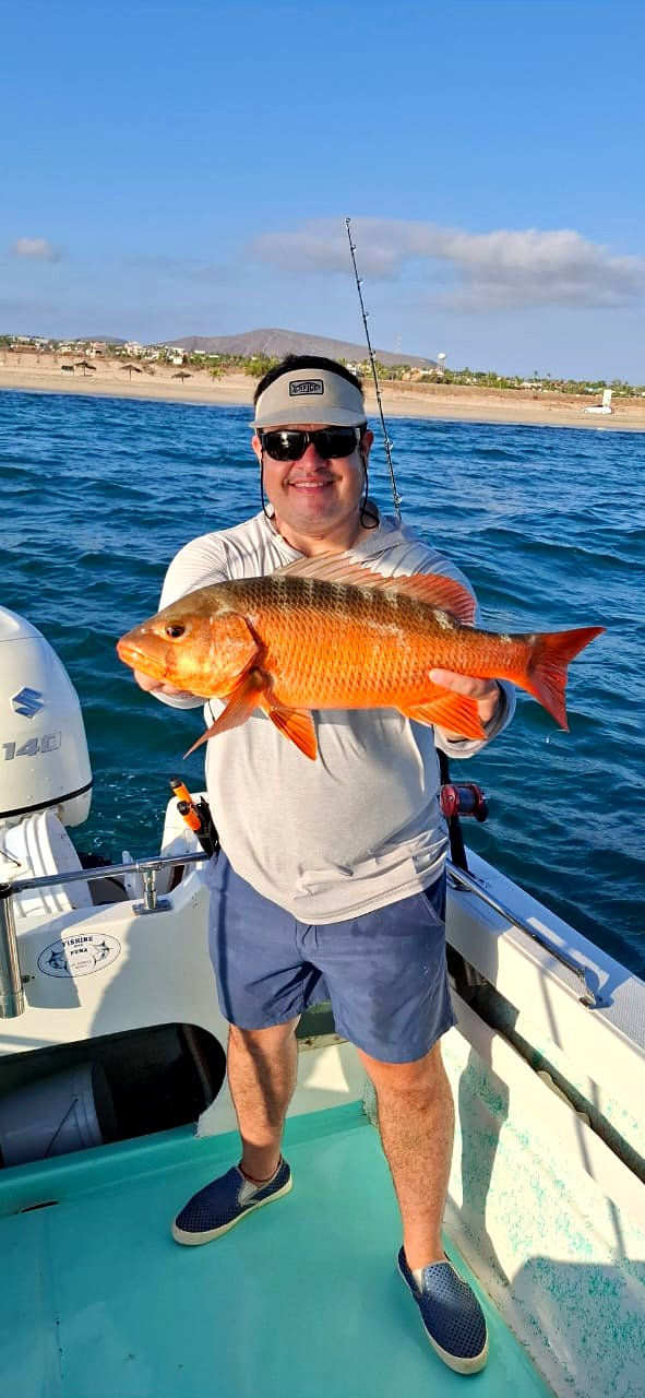 Smiling angler on a boat holding a red-orange barred snapper, with a sandy beach, palapas and a desert hill behind