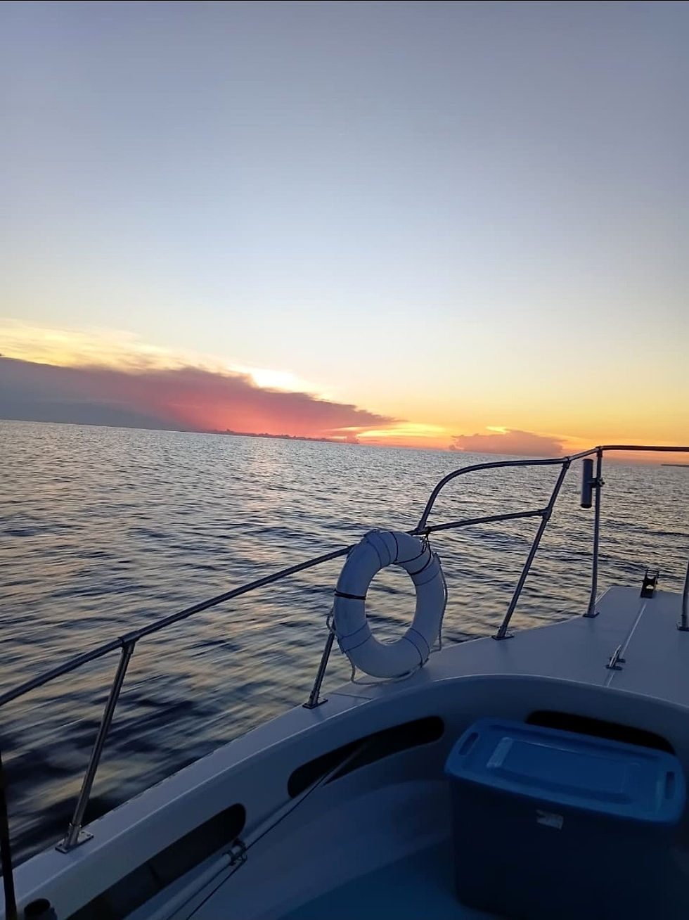 View from the bow of a boat over calm water at golden hour, a white life ring on the stainless rail, low pink and orange clouds on the horizon