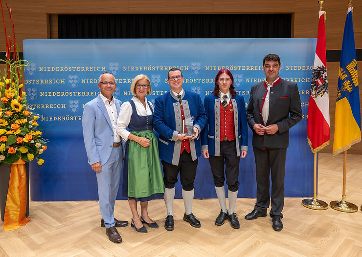 Fünf Personen vor blauem Banner mit „Niederösterreich“-Aufschrift, eine Frau im Dirndl, zwei Männer in Tracht, ein Mann im Anzug, eine junge Person in Tracht, ein Pokal.