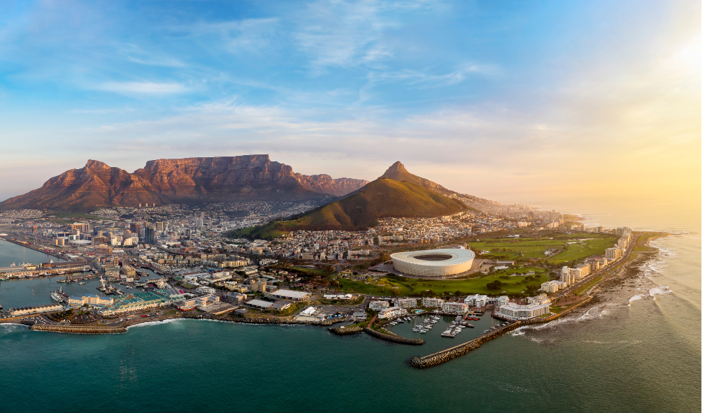 An aerial shot of Cape Town with Devil's Peak, Table Mountain, and Lion's Head, aswell as Atlantic seaboard and City Bowl pictured. The sun is coming up on the right hand side.