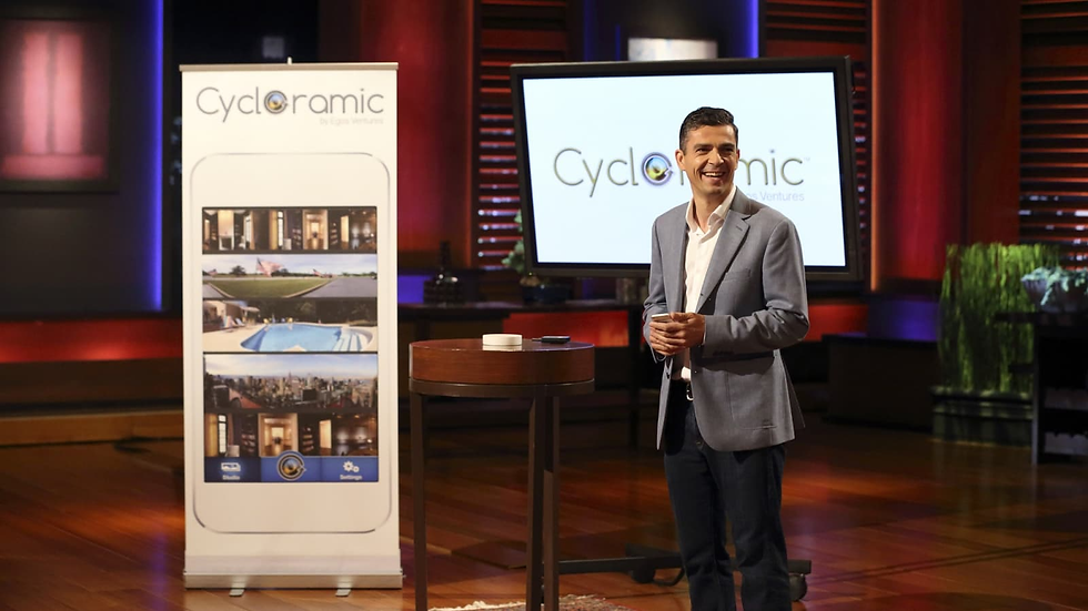 A man in a light blue blazer and dark jeans stands on the set of Shark Tank, smiling while presenting his product, Cycloramic. A large promotional banner next to him displays the Cycloramic app, which showcases panoramic photography features. A small round table in front of him holds a device. The background features the signature Shark Tank stage setup with a large screen displaying the Cycloramic logo.