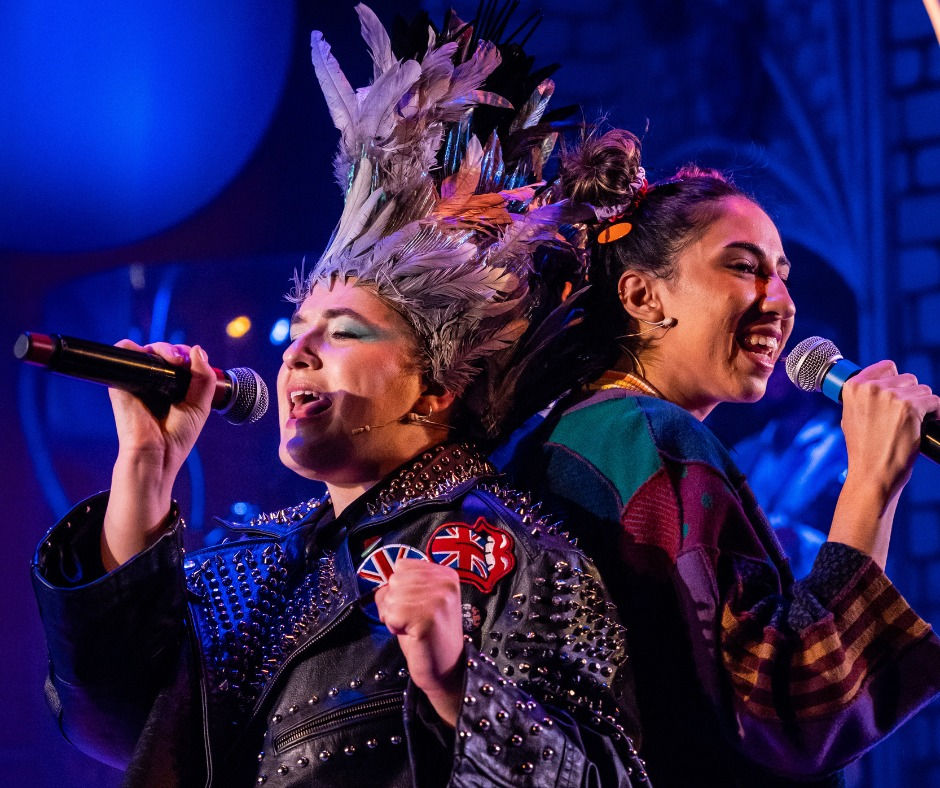 Two women standing back to back singing into hand held mics. One is dressed as The Pigeon, the other a young women wearing bunches and a cloth outfit