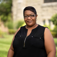 Smiling woman wearing glasses, black dress in front of a building. Hardon Education