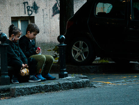 Two children sit on a curb, one holding a paper bag, the other with a toy. Graffiti-covered wall and parked car in the background.