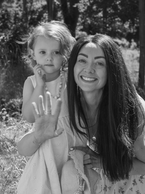 A black and white image of a mother with her young daughter, smiling and waving at the camera, surrounded by nature.
