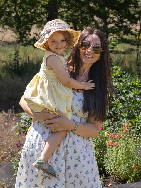 A mother holding her young daughter, smiling at the camera and surrounded by nature.