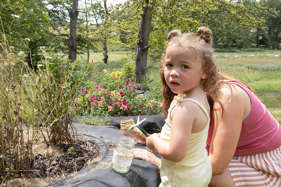 A little girl wearing a yellow top and space buns looking at the camera whilst snipping flowers from a Tyre Flowerbed with her mum next to her.