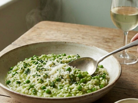A shallow ceramic bowl filled with bright green pea and mint risotto, topped with grated parmesan cheese, sitting on a rustic wooden table next to a glass of white wine in a Thomas Vaughan kitchen.